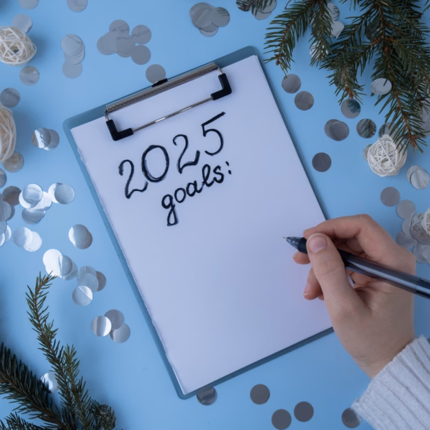 Widok z góry na dłoń piszącą na kartce na podkładce "2025 goals:" z ozdobami świątecznymi i konfetti wokół