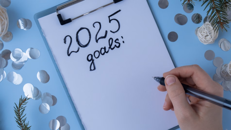 Widok z góry na dłoń piszącą na kartce na podkładce "2025 goals:" z ozdobami świątecznymi i konfetti wokół