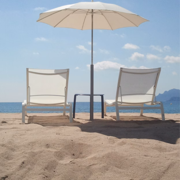 Dwa białe leżaki plażowe i mały stolik pod białym parasolem na piaszczystej plaży, z błękitnym morzem i niebem w tle.