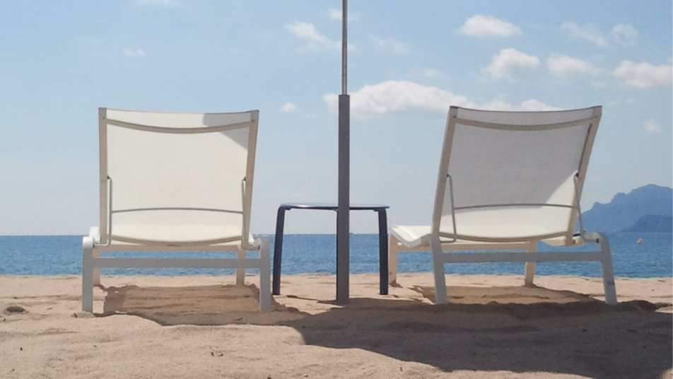 Dwa białe leżaki plażowe i mały stolik pod białym parasolem na piaszczystej plaży, z błękitnym morzem i niebem w tle.
