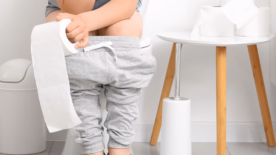 Dziecko w szarej koszulce i spodniach siedzące na toalecie, trzymające rolkę papieru toaletowego. Obok stoi kosz na śmieci i stolik z papierem toaletowym.