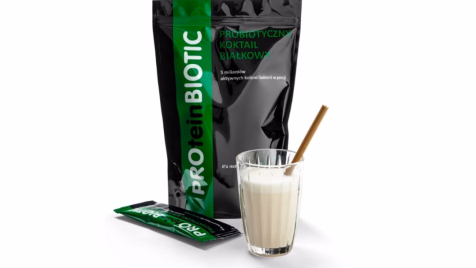 Opakowanie produktu PROteinBIOTIC, obok szklanka z białym koktajlem i słomką, a także mała, pojedyncza saszetka produktu