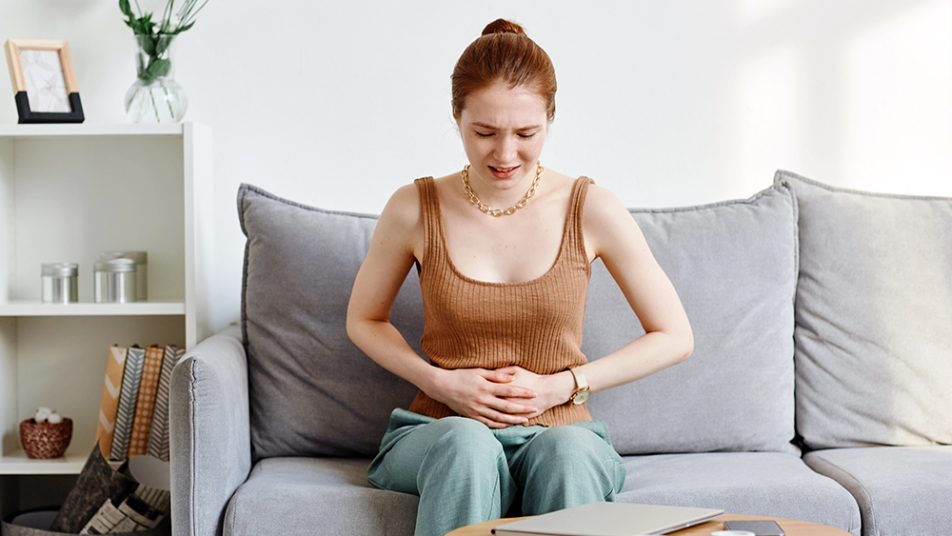 Kobieta siedzi na kanapie i trzyma się za brzuch z bolesnym wyrazem twarzy.