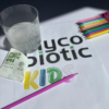 szklanka z rozpuszczonym produktem Mycobiotic KIDS w tle kolorowe kredki i opakowanie produktu2