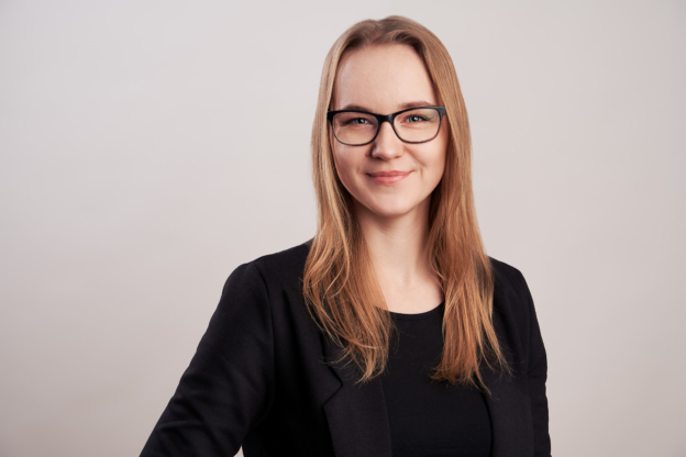 Profesjonalne zdjęcie portretowe młodej kobiety w okularach, uśmiechniętej, ubranej w czarną marynarkę i czarną bluzkę na jasnym, neutralnym tle